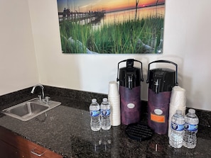 A kitchen counter with a sink, water bottles, paper cups, and two water dispensers. - Studio 6 Suites Jacksonville, NC (Jacksonville)