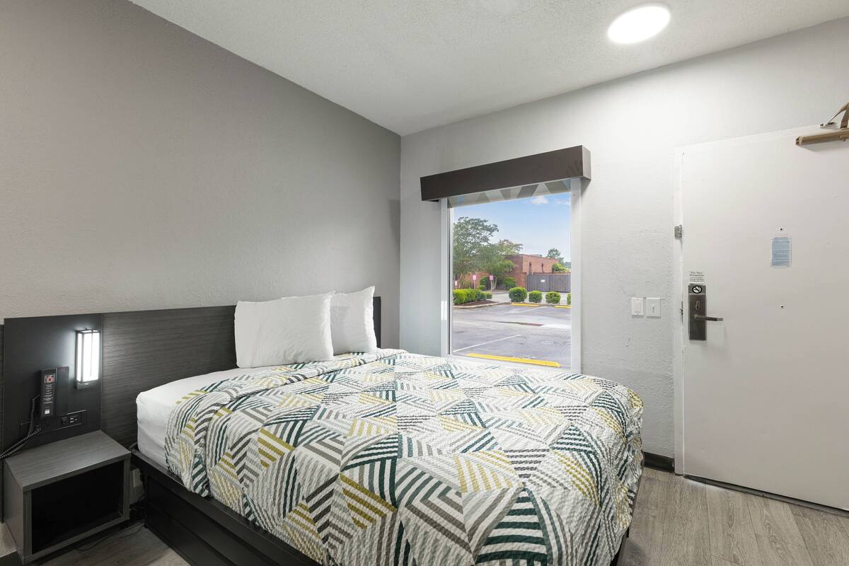A hotel room with a bed, a nightstand, a window, and a door.