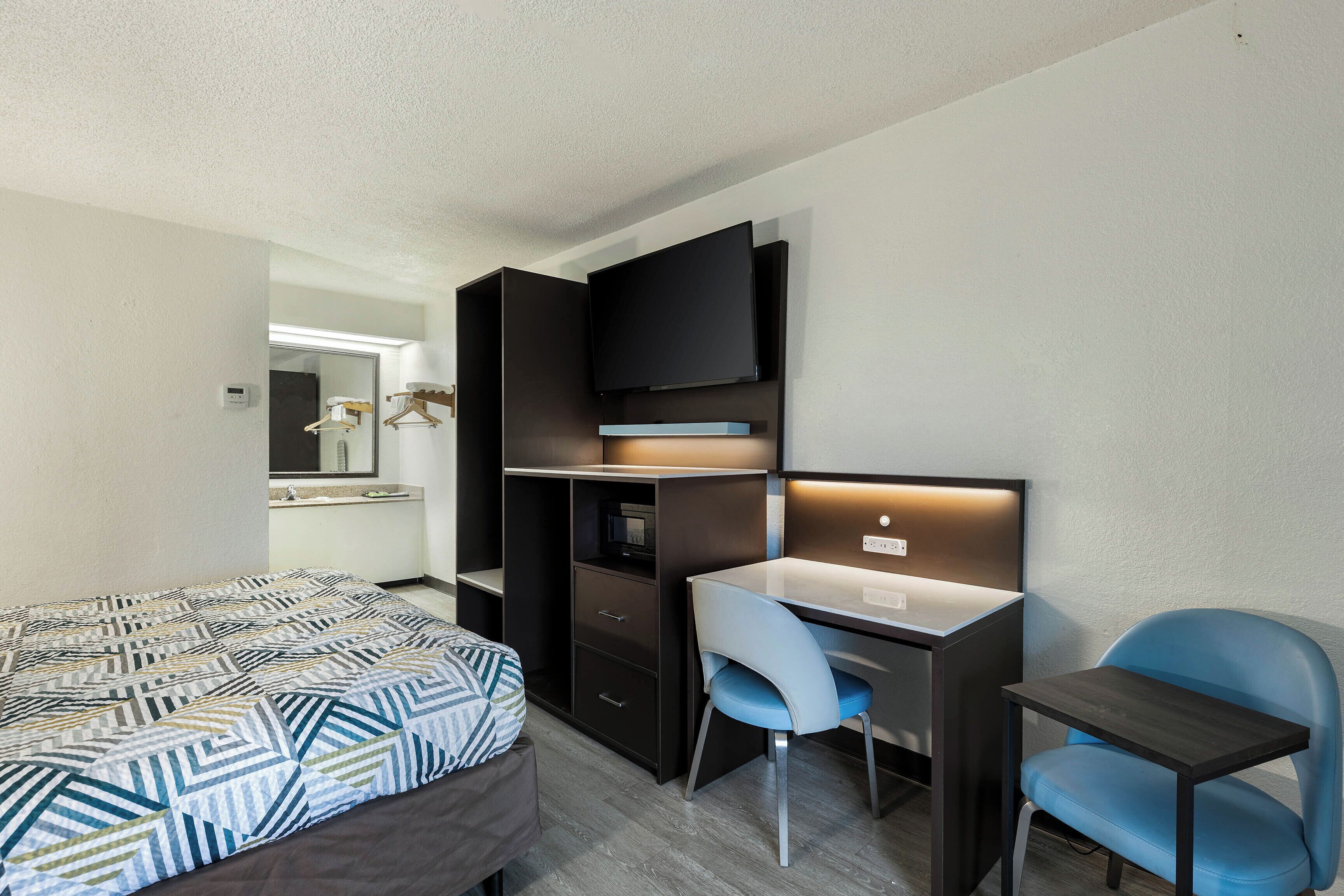 A compact hotel room with a bed, a desk, a chair, a TV, and a mirror.
