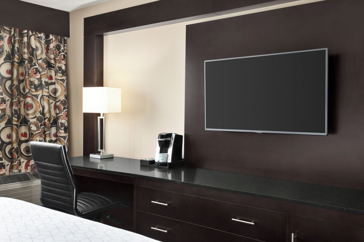A hotel room with a bed, a desk with a lamp, a coffee maker, and a TV on the wall.