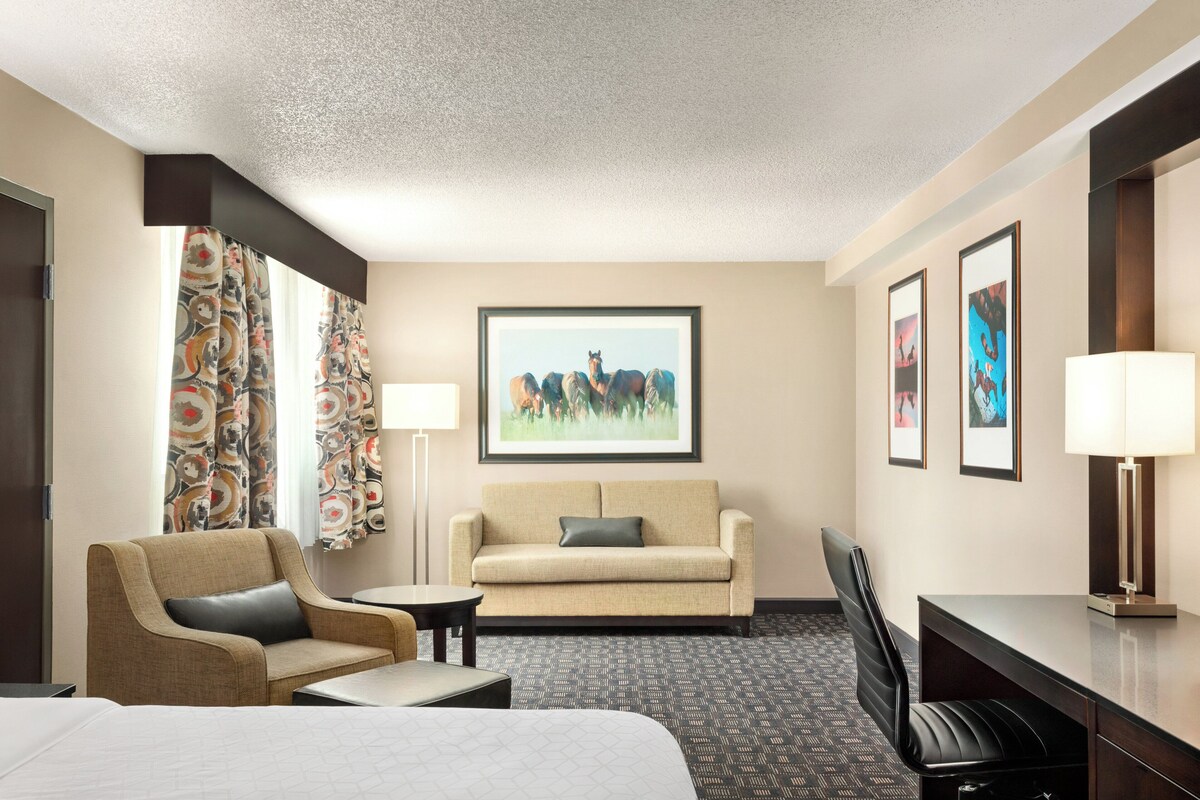 A hotel room with a bed, a sofa, a desk, and a chair. There are two lamps, a window with curtains, and a framed picture on the wall.