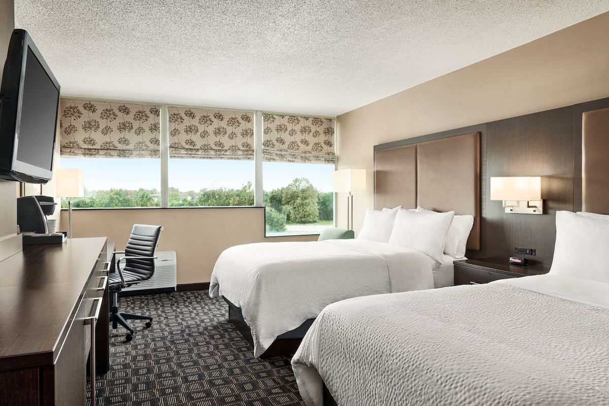 A hotel room with two beds, a desk, a chair, a TV, and a window with a view of trees.