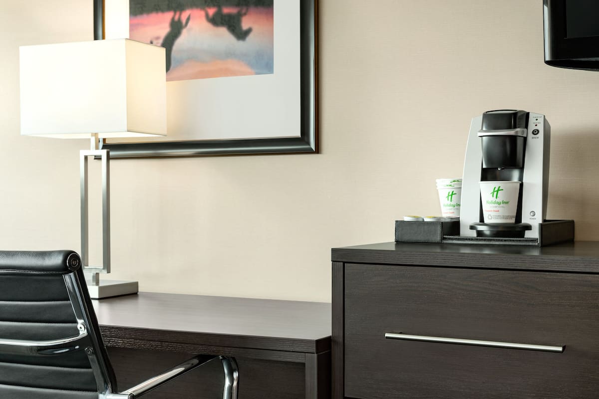 A hotel room with a desk, a coffee maker, a framed picture, and a lamp.
