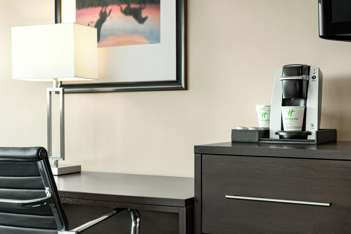 A hotel room with a desk, a coffee maker, a framed picture, and a lamp.
