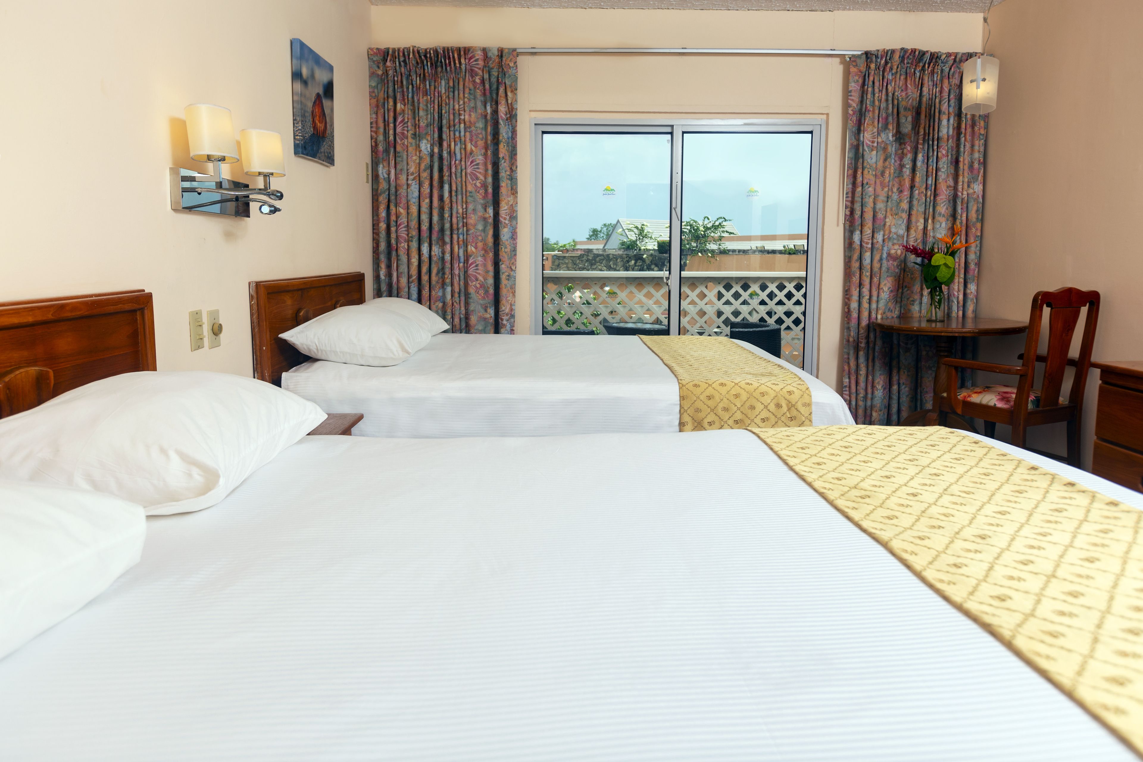 Ein Hotelzimmer mit zwei Betten, einem Balkon mit Aussicht, einem kleinen Tisch mit Stuhl und einer Wandleuchte.