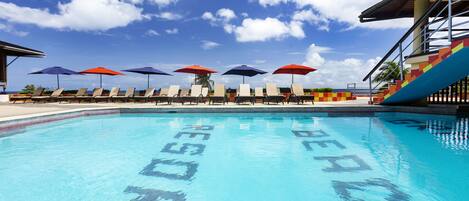 Una piscina con sillones de descanso y sombrillas, cielo despejado y un edificio con una escalera de colores.