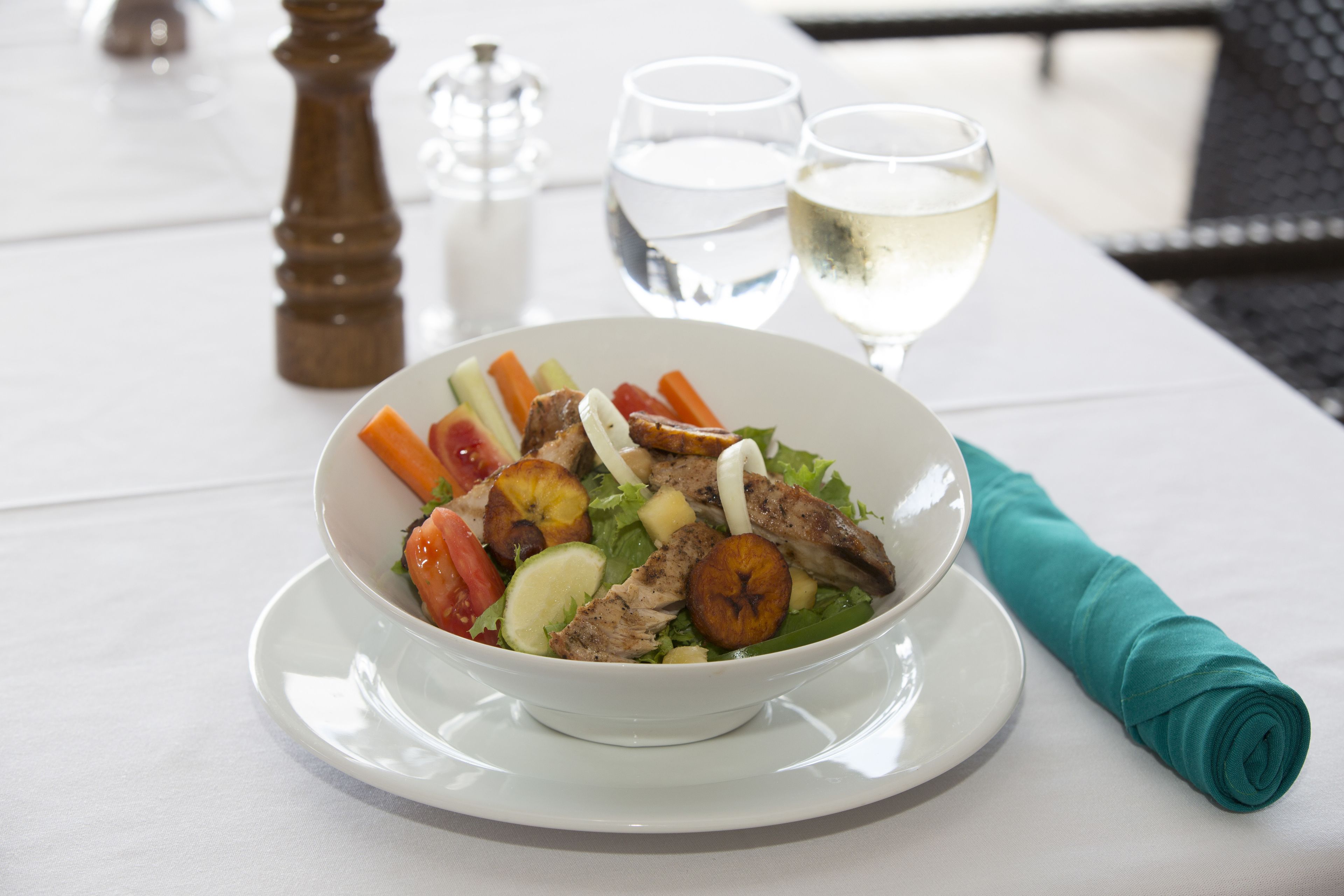Une table blanche avec un saladier rempli de salade, deux verres de vin blanc et une serviette verte roulée.