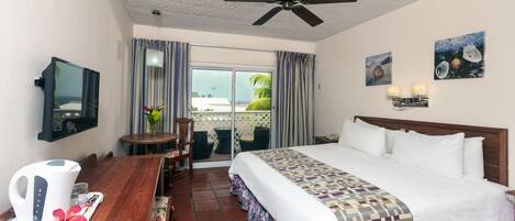 A hotel room with a bed, a desk with a kettle, a chair, a ceiling fan, and a view through the window.