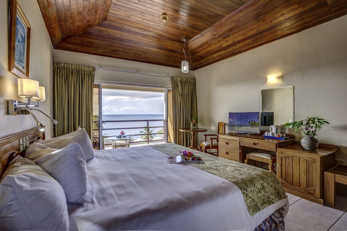 A hotel room with a large bed, wooden ceiling, a desk with a computer, a TV, and a balcony with a view of the ocean.
