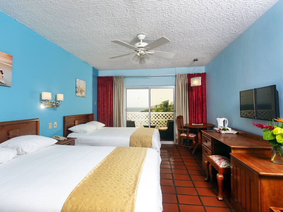 A hotel room with two beds, a desk, a chair, a television, and a balcony with red curtains.