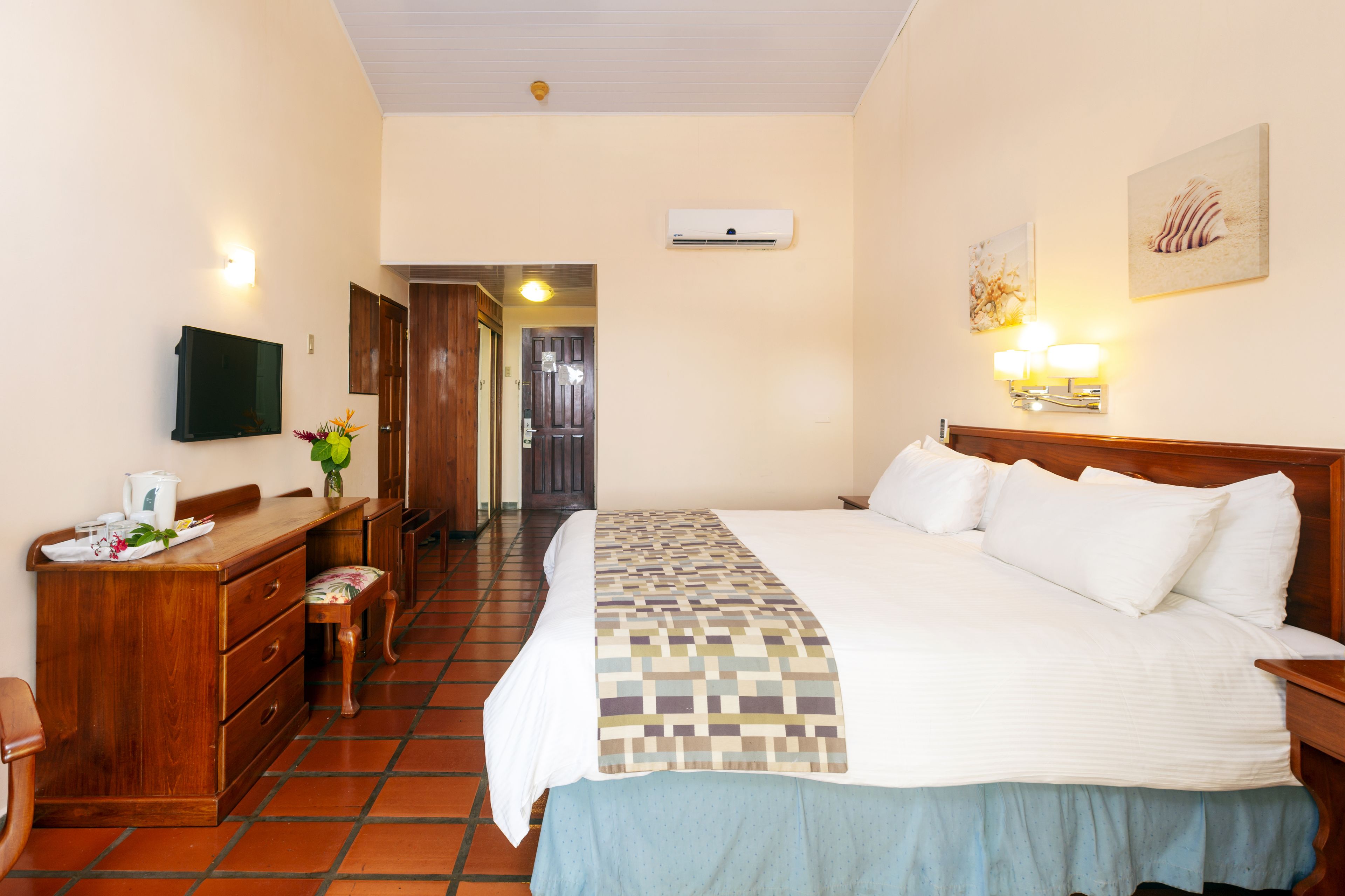 A hotel room with a bed, a desk, a chair, a TV, a wall-mounted air conditioner, and a door leading to another room.