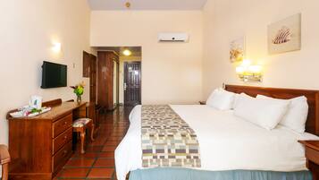 Habitación de hotel con cama, escritorio, silla, televisor, aire acondicionado instalado en la pared y puerta que da a otra habitación.