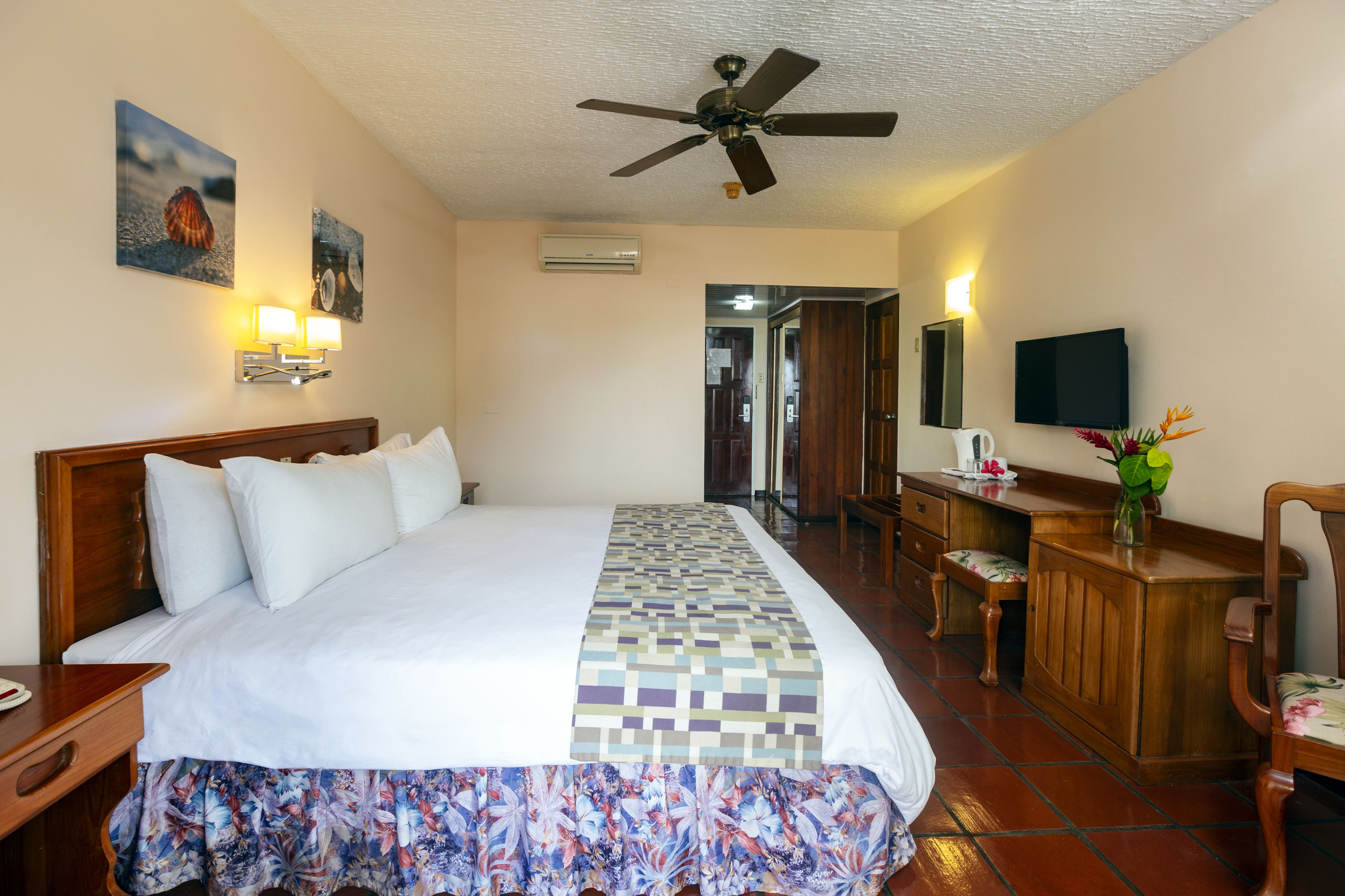 A hotel room with a bed, a desk, a chair, a ceiling fan, and a television.