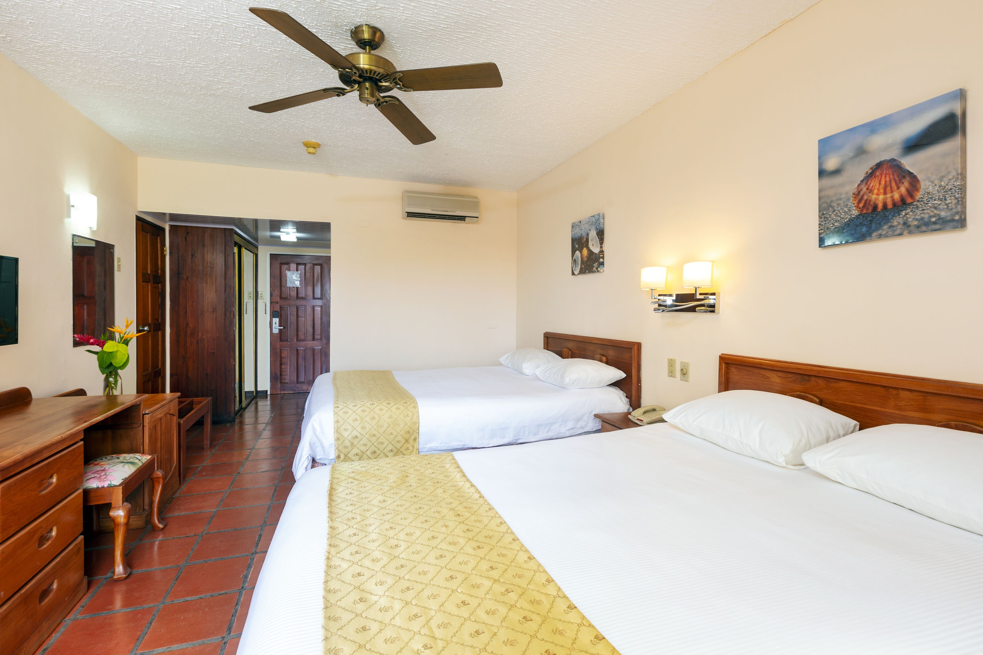 Habitación de hotel con dos camas, un escritorio, una silla, un ventilador de techo y un cuadro en la pared.