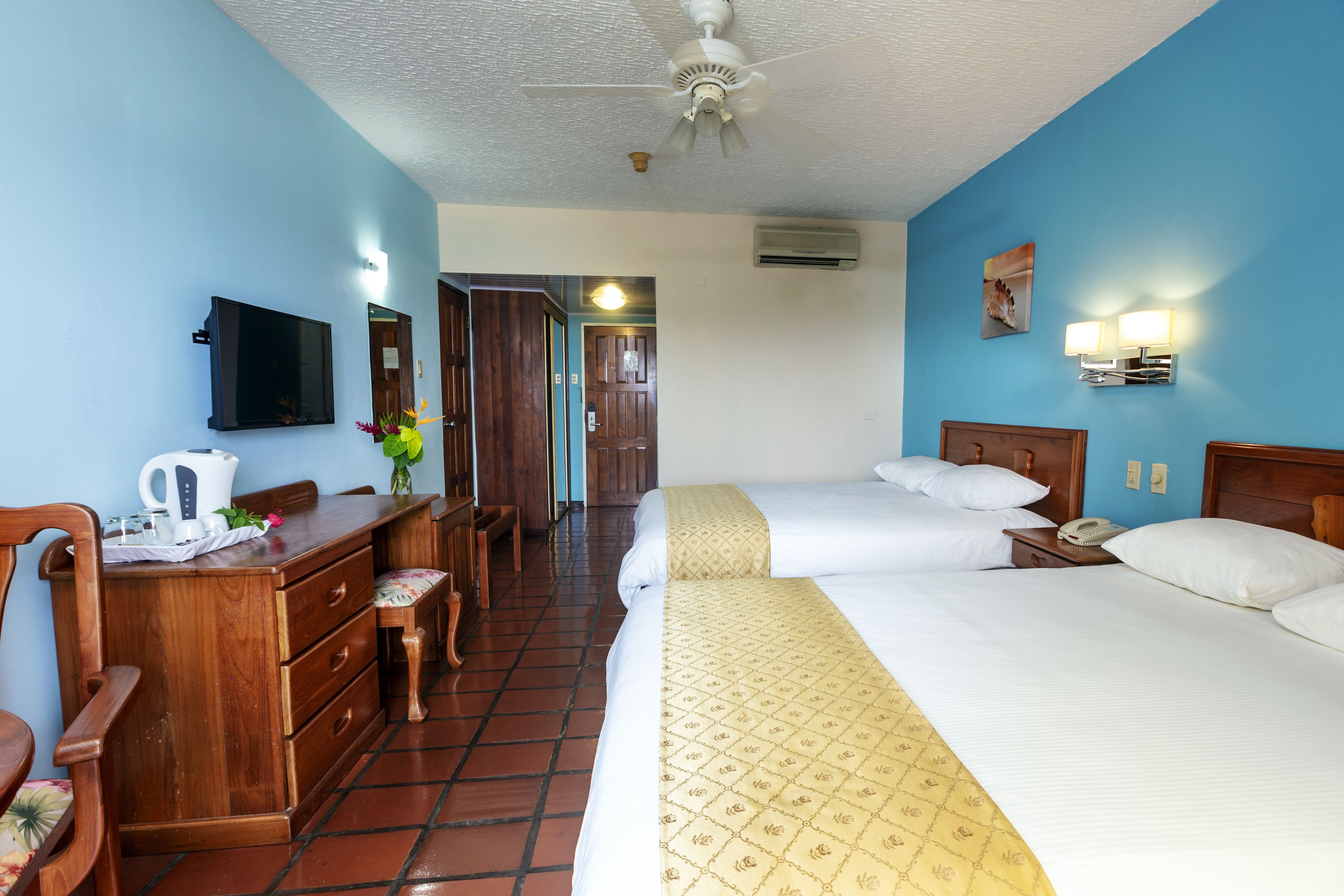 A hotel room with two beds, a desk with a chair, a television, and a ceiling fan.