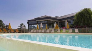 Una piscina con sillas de descanso y sombrillas, un edificio moderno con amplias ventanas y un cielo despejado.