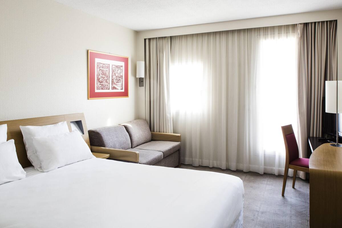Ein Hotelzimmer mit Bett, einer Couch, einem Schreibtisch mit Stuhl, einem gerahmten Bild an der Wand und einem Fenster mit Vorhängen.