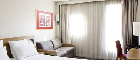 Habitación de hotel con cama, sofá, escritorio y silla, un cuadro en la pared y ventana con cortinas.