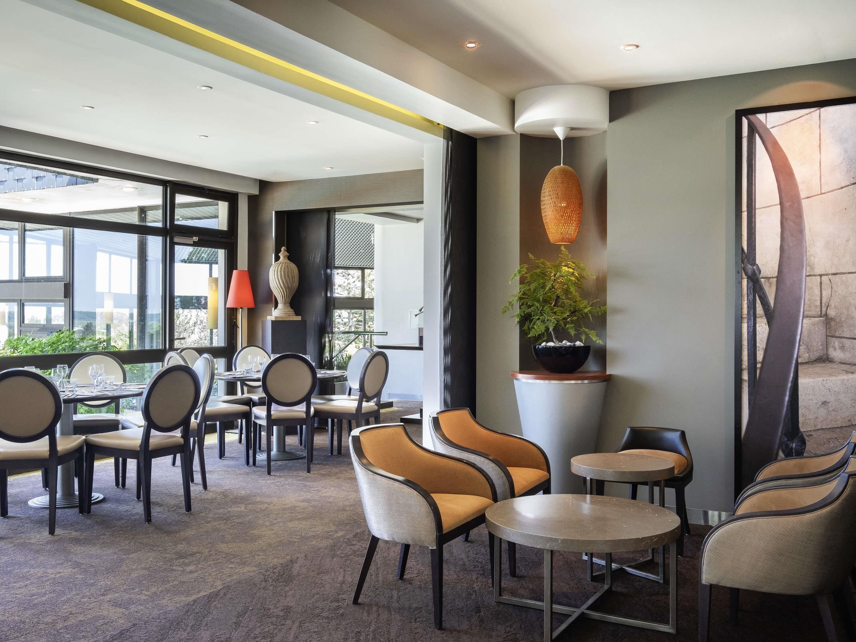 A modern hotel lobby with round tables and chairs, a large window offering a view, and a decorative wall with a plant.