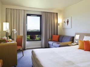Habitación de hotel con cama, sofá, escritorio con lámpara, ventana con cortinas y un cuadro en la pared.