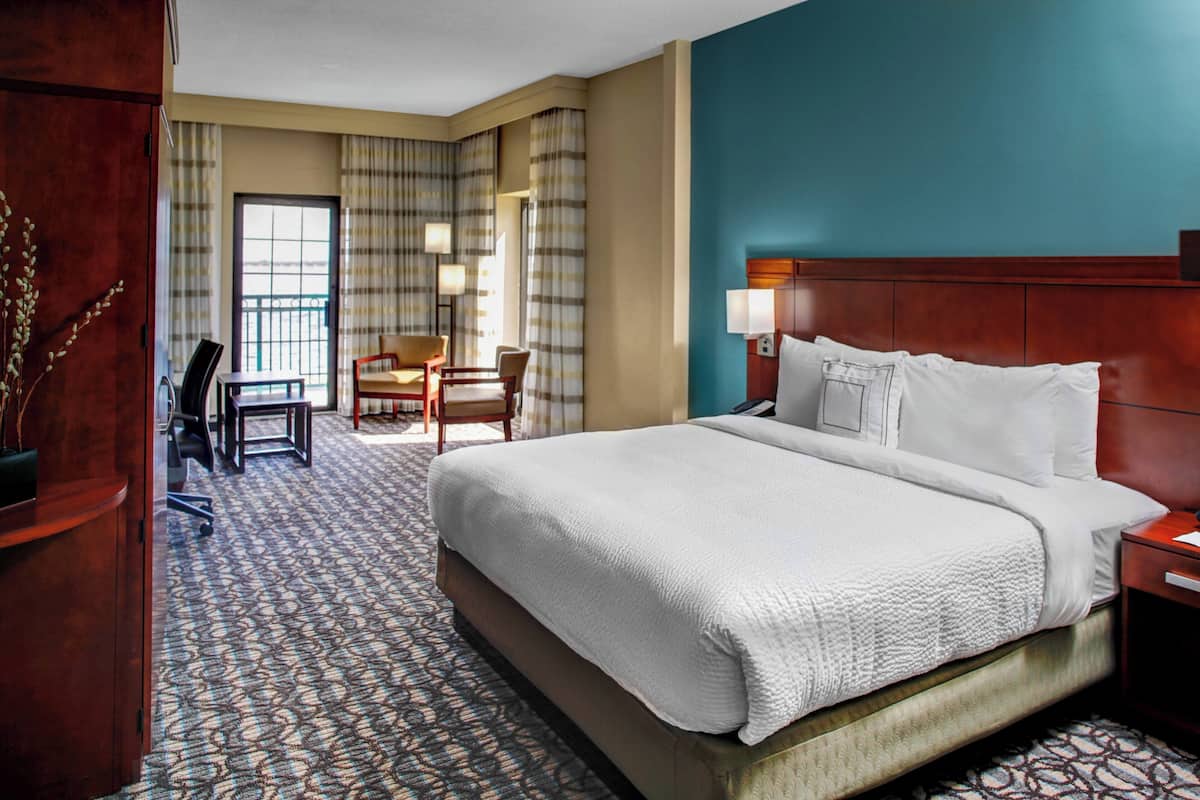 A hotel room with a large bed, a desk with a chair, a small table, and a view through a window with curtains.