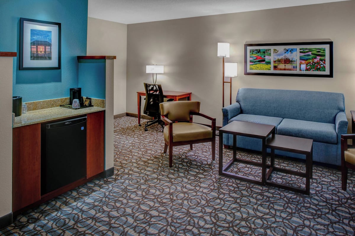 A hotel room with a kitchenette, a blue sofa, a small table, a chair, and a patterned carpet.