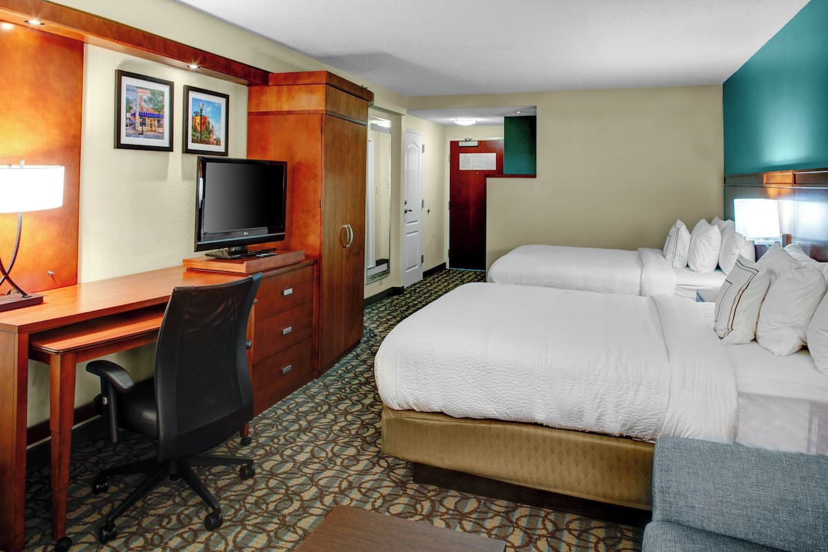 A hotel room with a bed, a desk with a computer, a TV, and a chair. There is a door leading to another room.