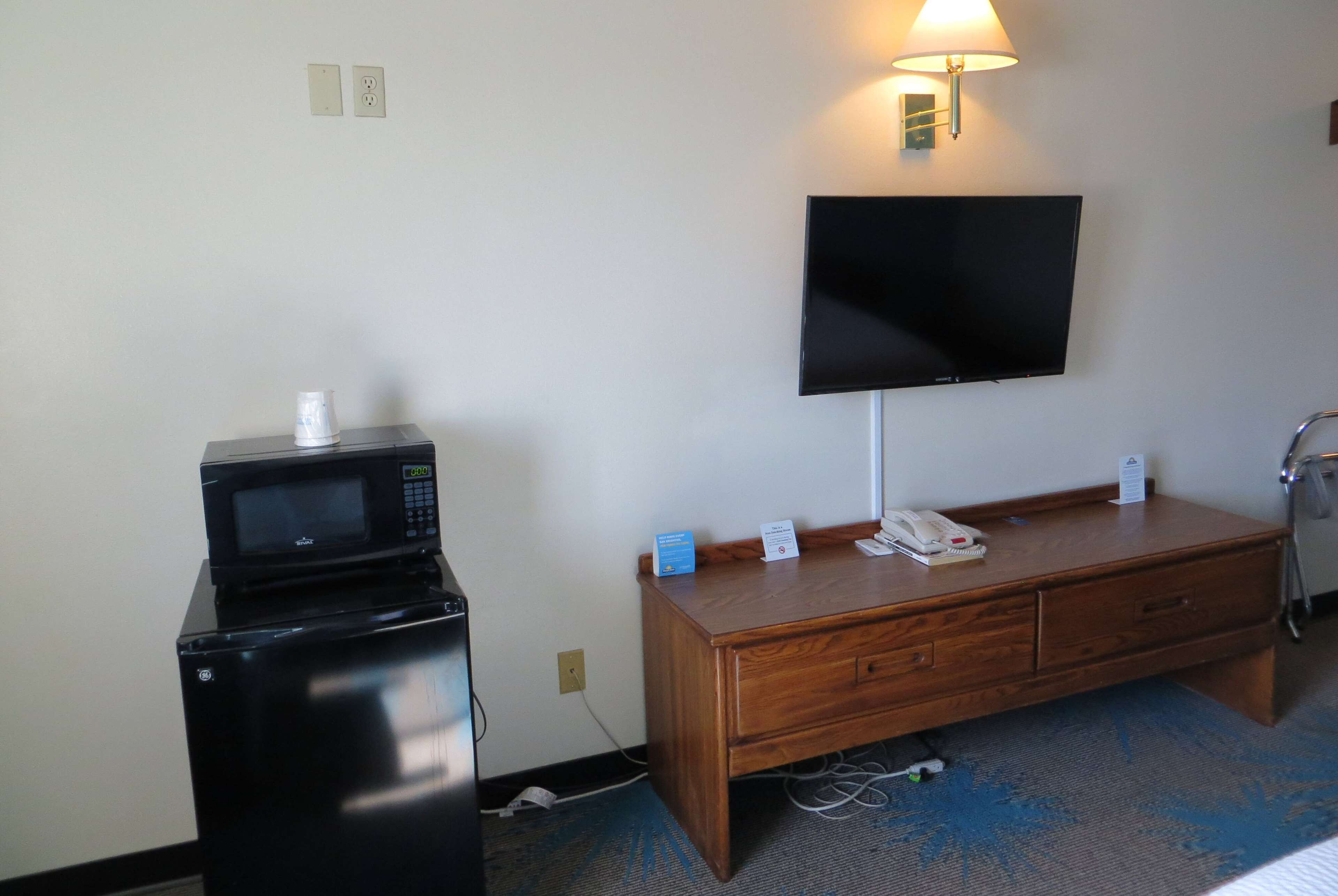 Habitación de hotel con televisor de pantalla plana en la pared, un escritorio de madera con teléfono y un cartel, un microondas sobre un mueble, y una lámpara en el escritorio.