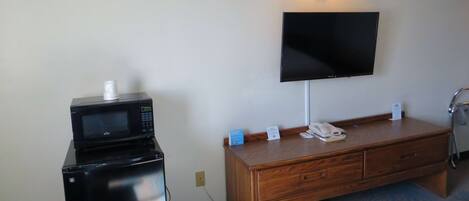 Habitación de hotel con televisor de pantalla plana en la pared, un escritorio de madera con teléfono y un cartel, un microondas sobre un mueble, y una lámpara en el escritorio.