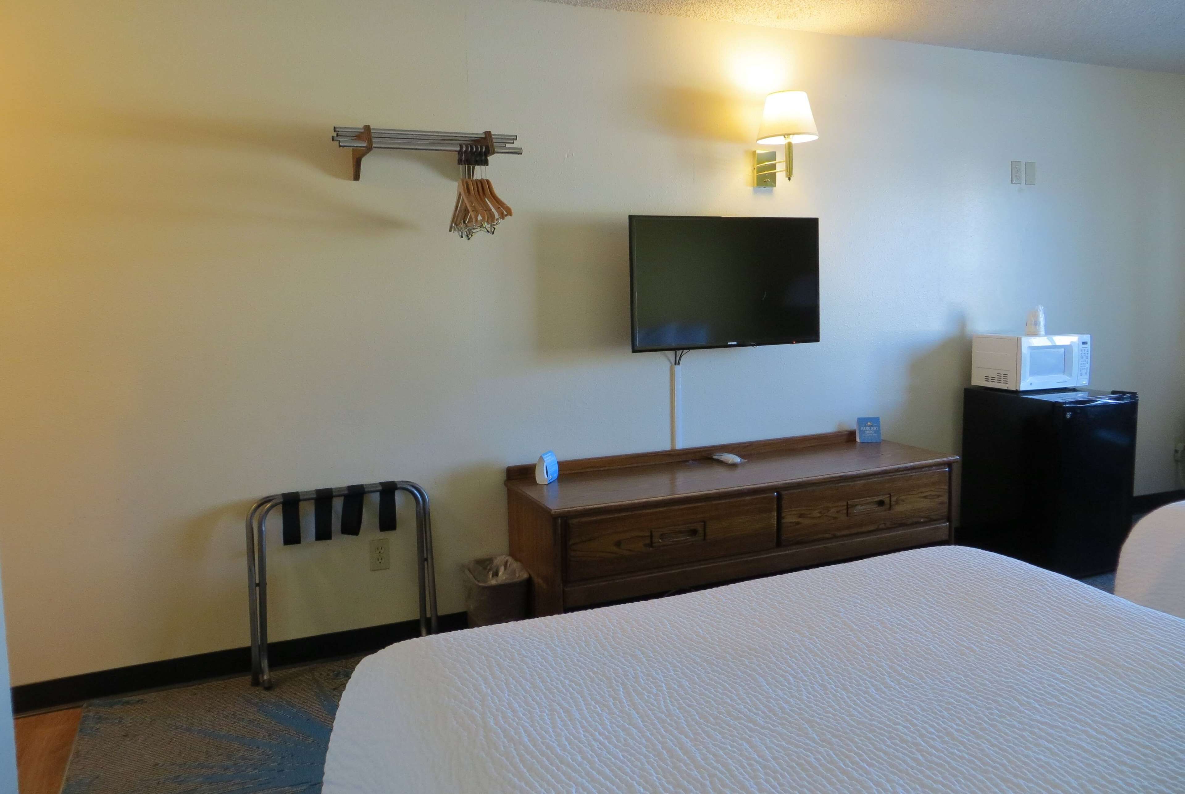 Habitación de hotel con cama, televisión montada en la pared, cómoda de madera, microondas y cubo de basura.