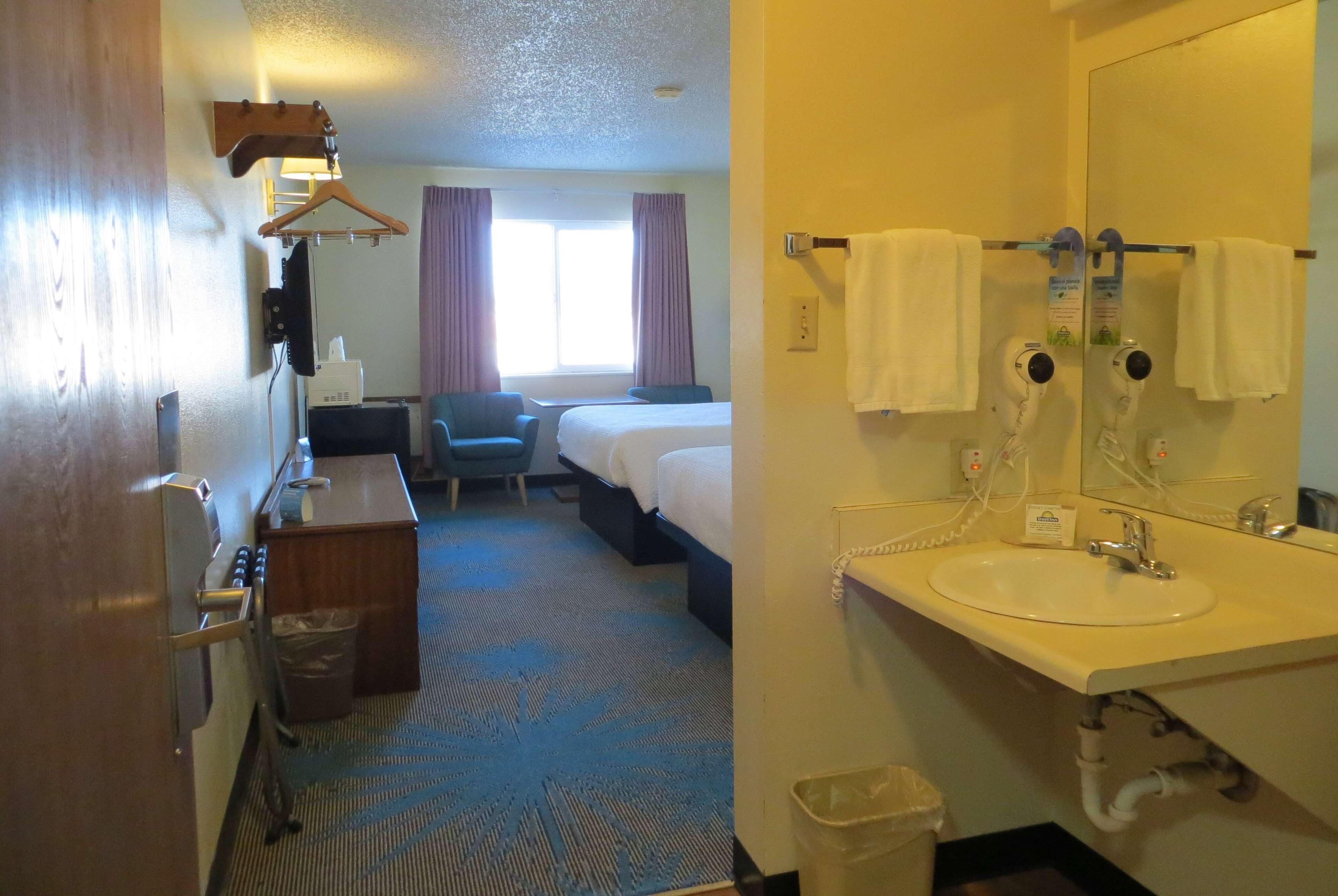 Habitación de hotel con cama, escritorio, silla, baño con lavamanos y espejo, y ventana con cortinas.