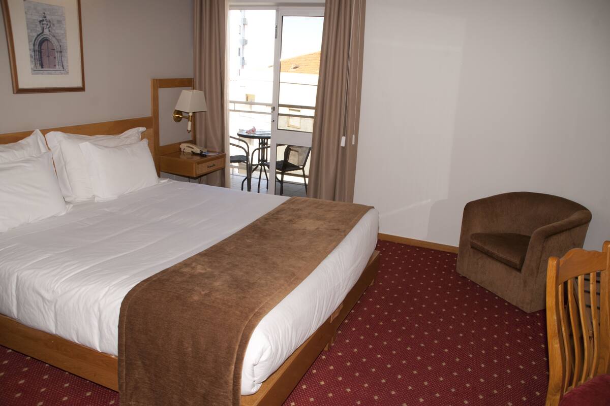 Habitación de hotel con una cama grande, una mesita de noche de madera con una lámpara, una silla y vistas al balcón exterior.