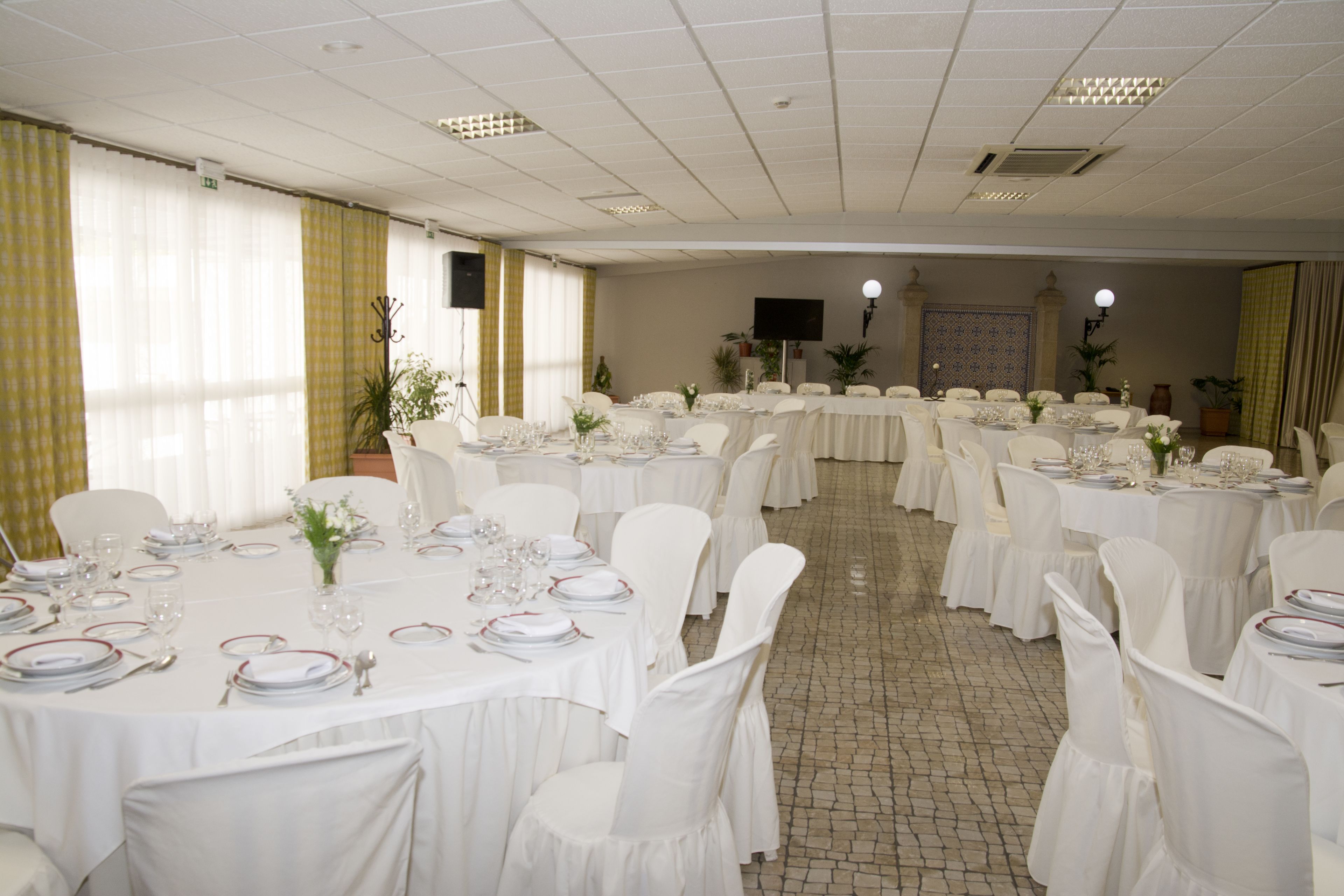 Een feestzaal met meerdere ronde tafels gedekt voor een diner, witte stoelen en een betegelde vloer.