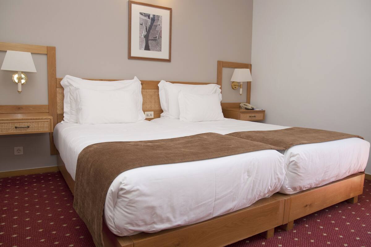 Cama doble con ropa de cama blanca y una manta marrón, cabecera de madera, lámparas de noche y un cuadro en la pared.