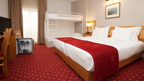Habitación de hotel con literas, un escritorio con silla, televisor, minibar y piso cubierto con alfombra roja.