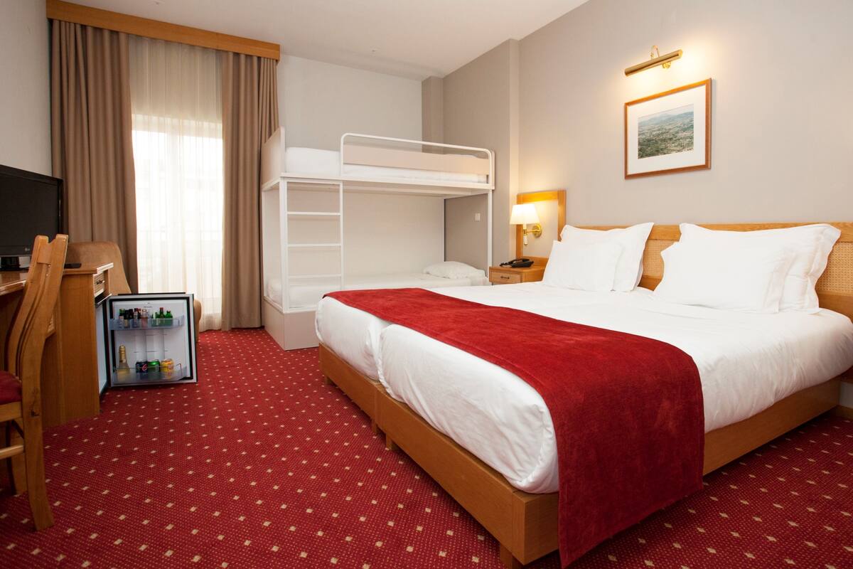 Habitación de hotel con literas, un escritorio con silla, televisor, minibar y piso cubierto con alfombra roja.