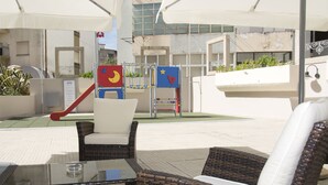 Un área de estar al aire libre con muebles de mimbre y una mesa de vidrio, un parque infantil con juegos de colores, y un edificio de fachada blanca al fondo.