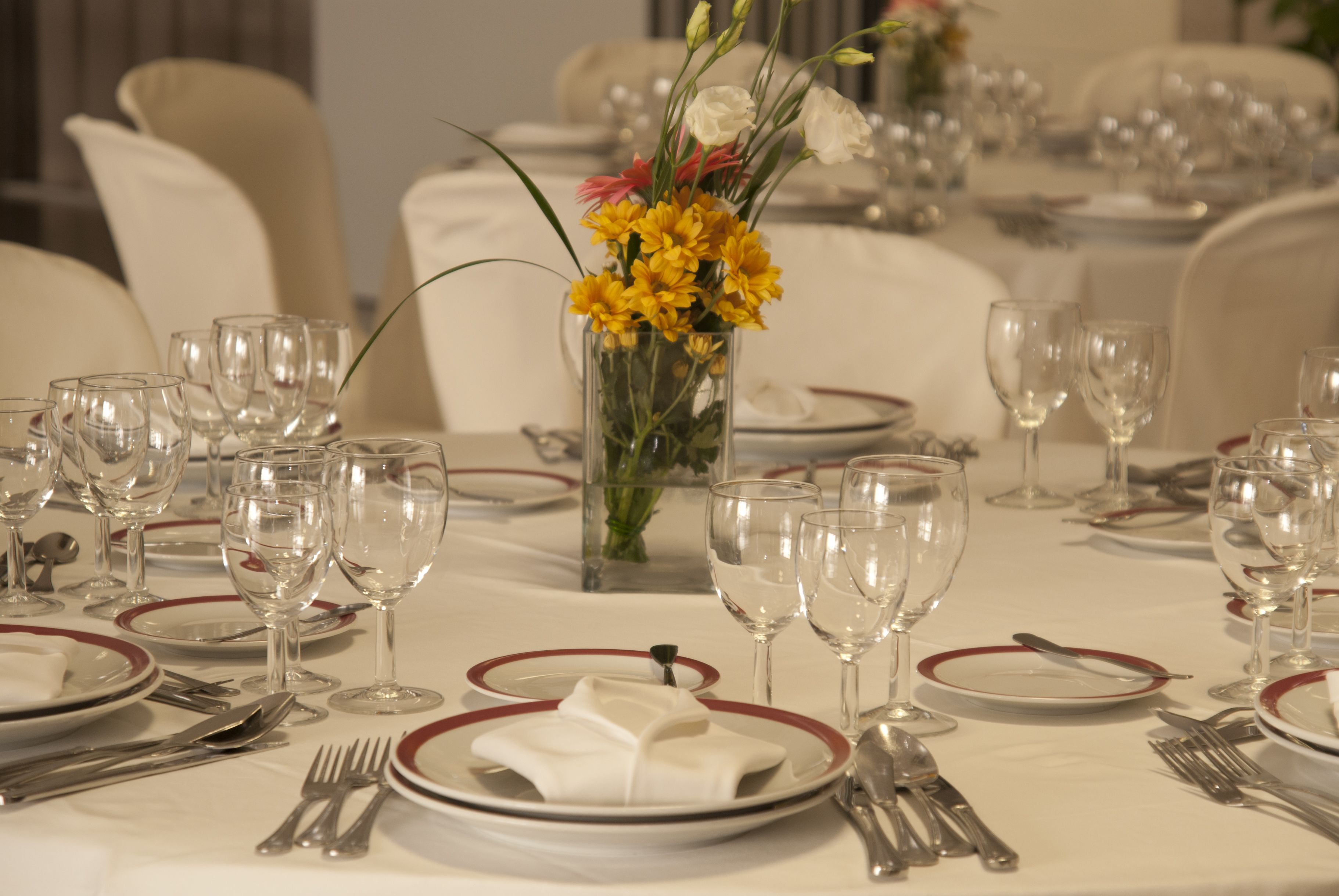 Une table à manger dressée avec de nombreux verres à vin, des assiettes et des couverts, agrémentée d’un centre de table composé de fleurs jaunes.