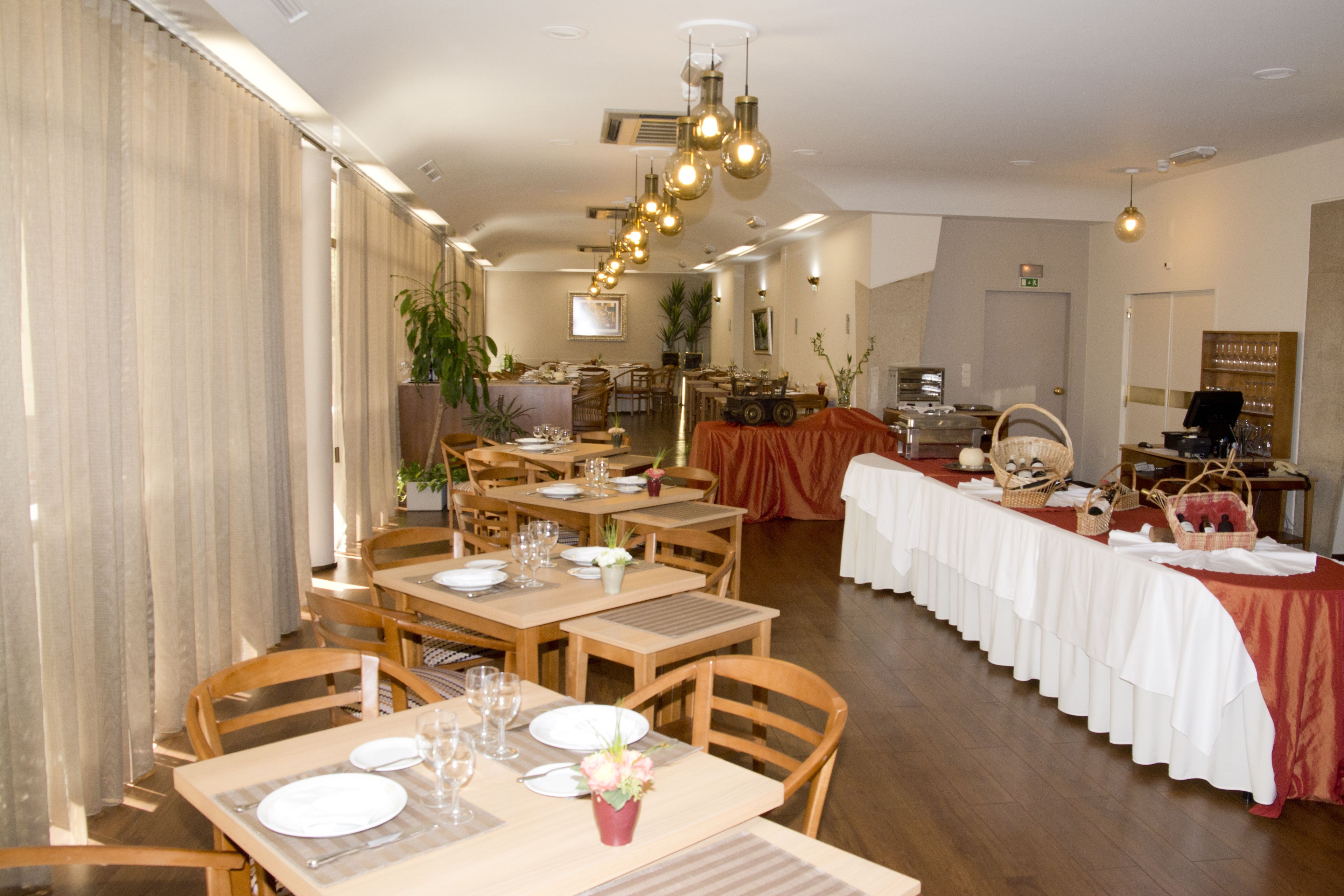 A restaurant with wooden tables and chairs, a long counter with a red tablecloth, and hanging light fixtures.