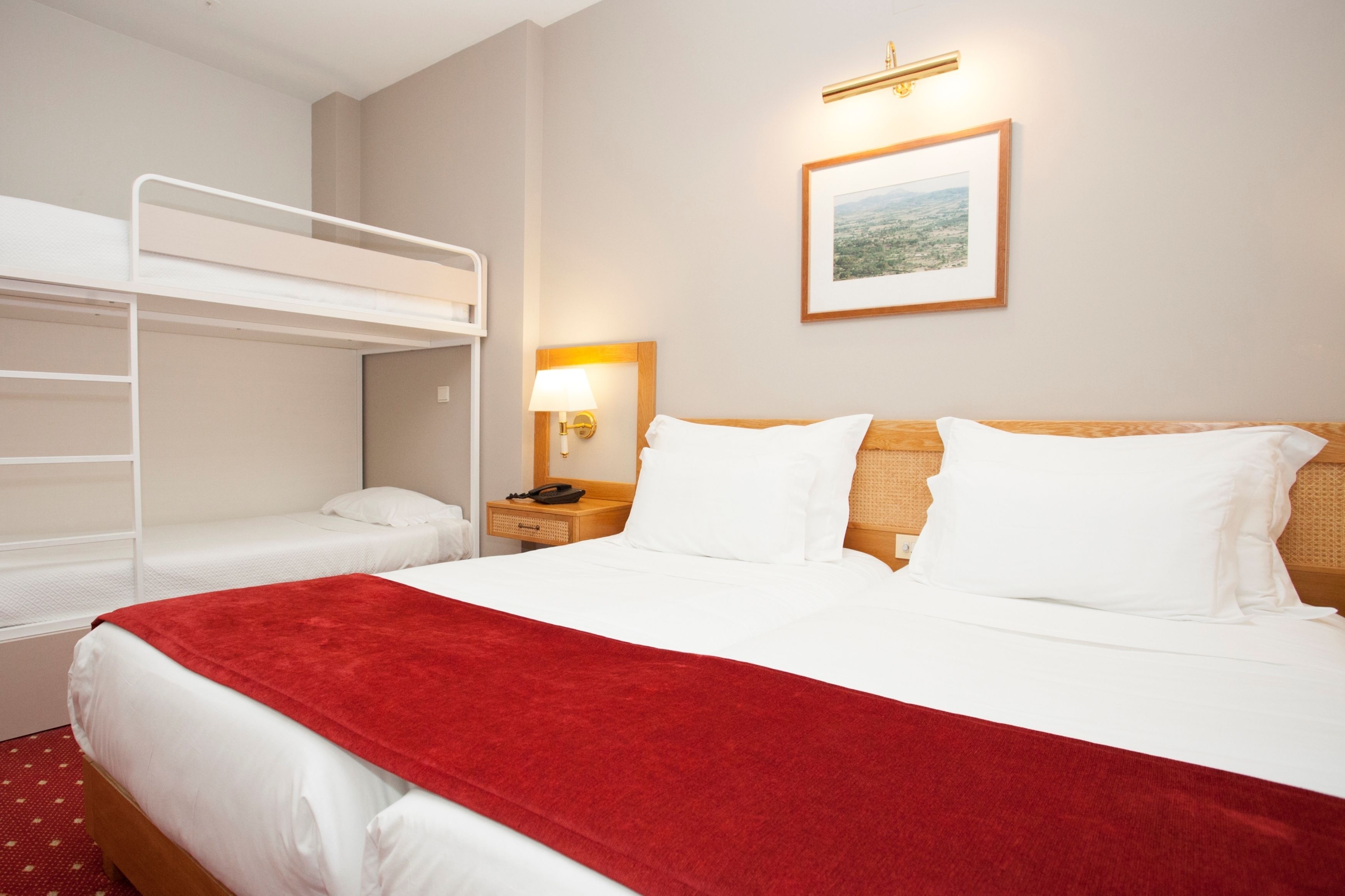 A hotel room with a bunk bed, a single bed, a red throw blanket, a bedside lamp, and a framed picture on the wall.