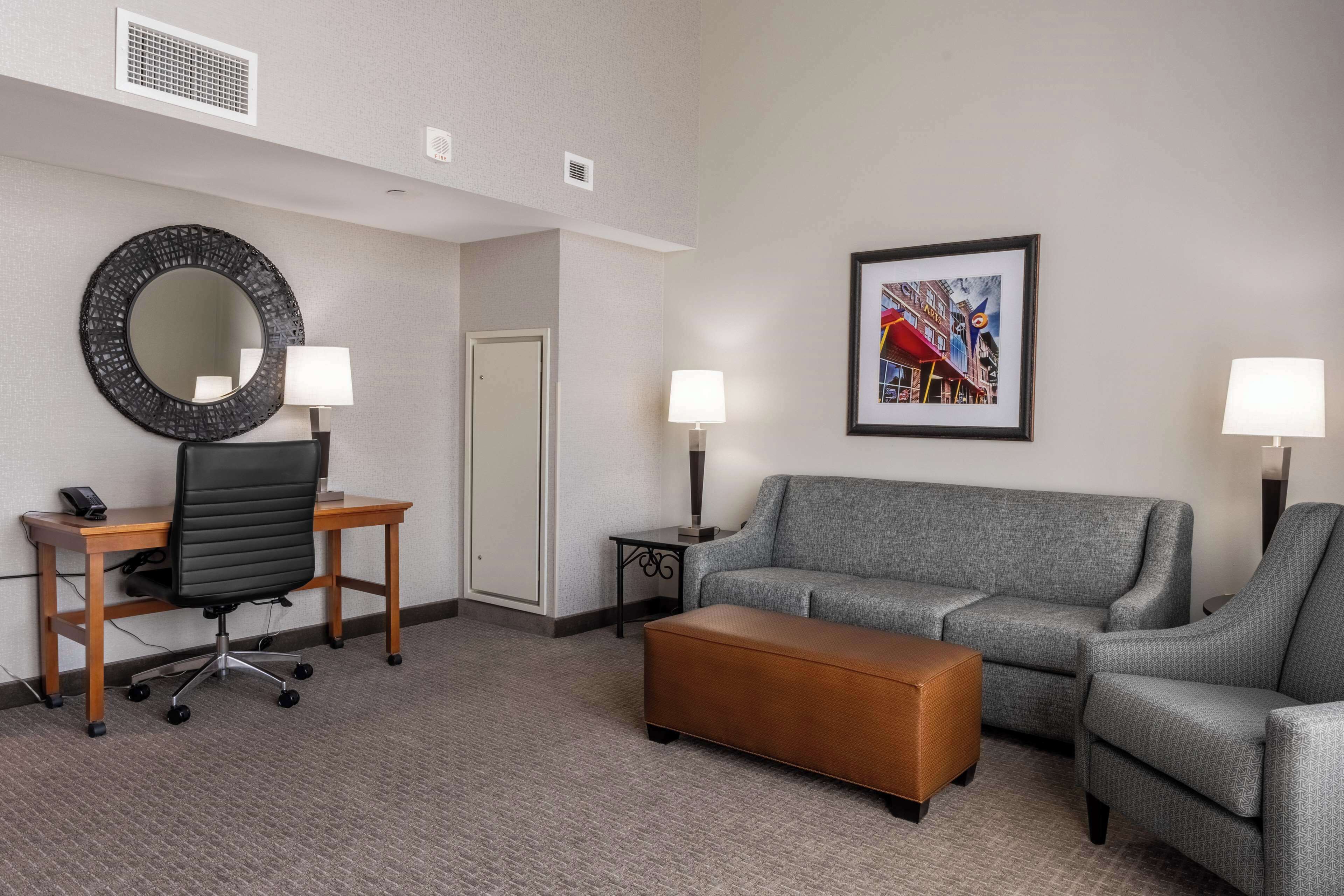 Une chambre d’hôtel avec un canapé gris, un bureau en bois avec une chaise, un miroir rond, un tableau encadré au mur et deux lampadaires.