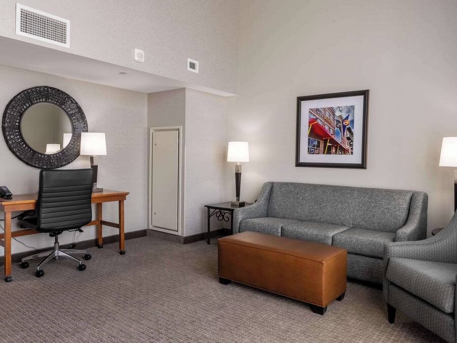 Une chambre d’hôtel avec un canapé gris, un bureau en bois avec une chaise, un miroir rond, un tableau encadré au mur et deux lampadaires.