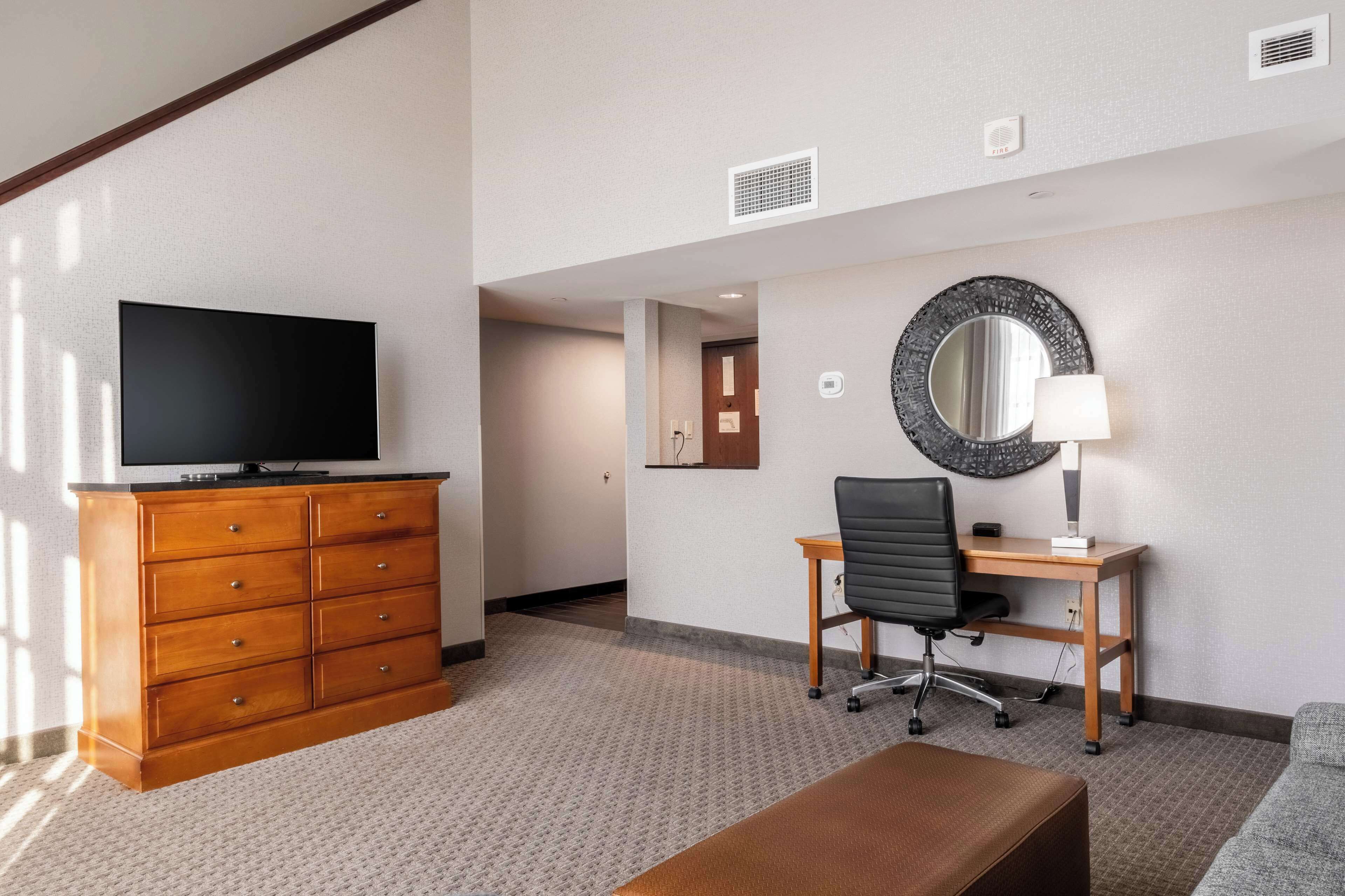 A hotel room with a wooden dresser, a flat-screen TV, a desk with a chair, a round mirror, and a lamp.