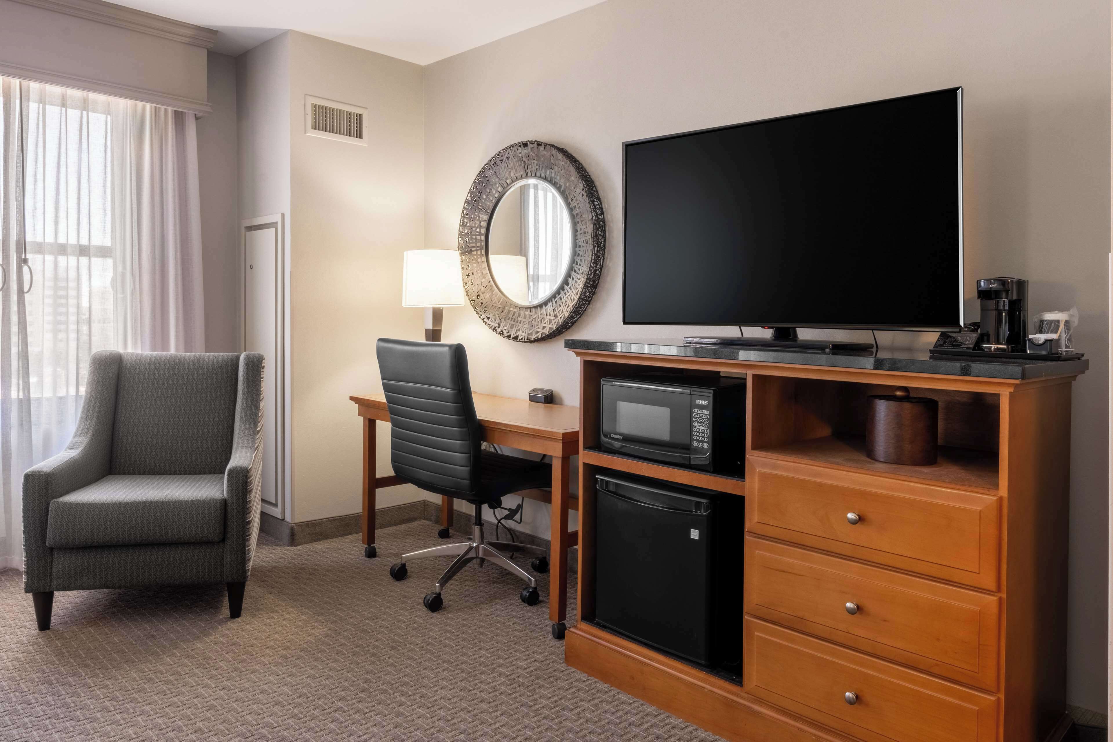 A hotel room with a flat-screen TV, a microwave, a small refrigerator, a desk with a chair, and a large mirror on the wall.