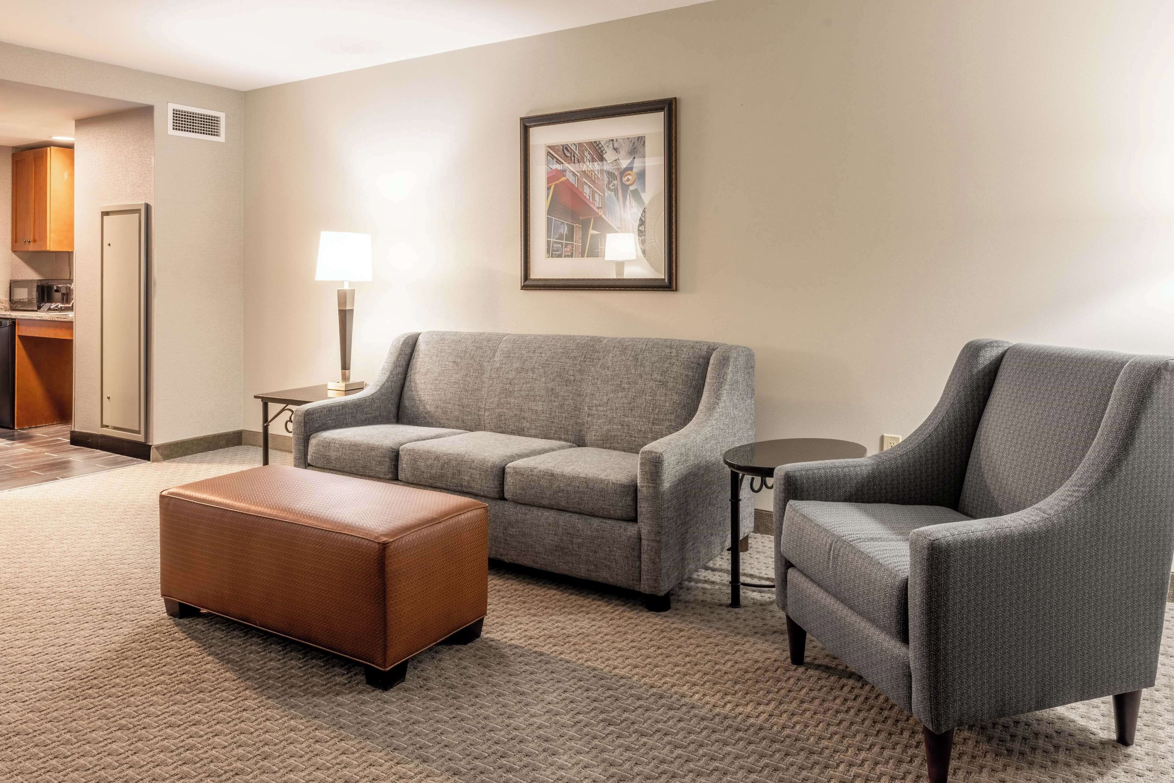 A living room with a grey sofa, a brown ottoman, and a grey armchair. There is a small table with a lamp, a framed picture on the wall, and a kitchenette in the background.