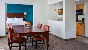 A hotel room with a bed, a dining table with chairs, a kitchenette, and a blue accent wall.