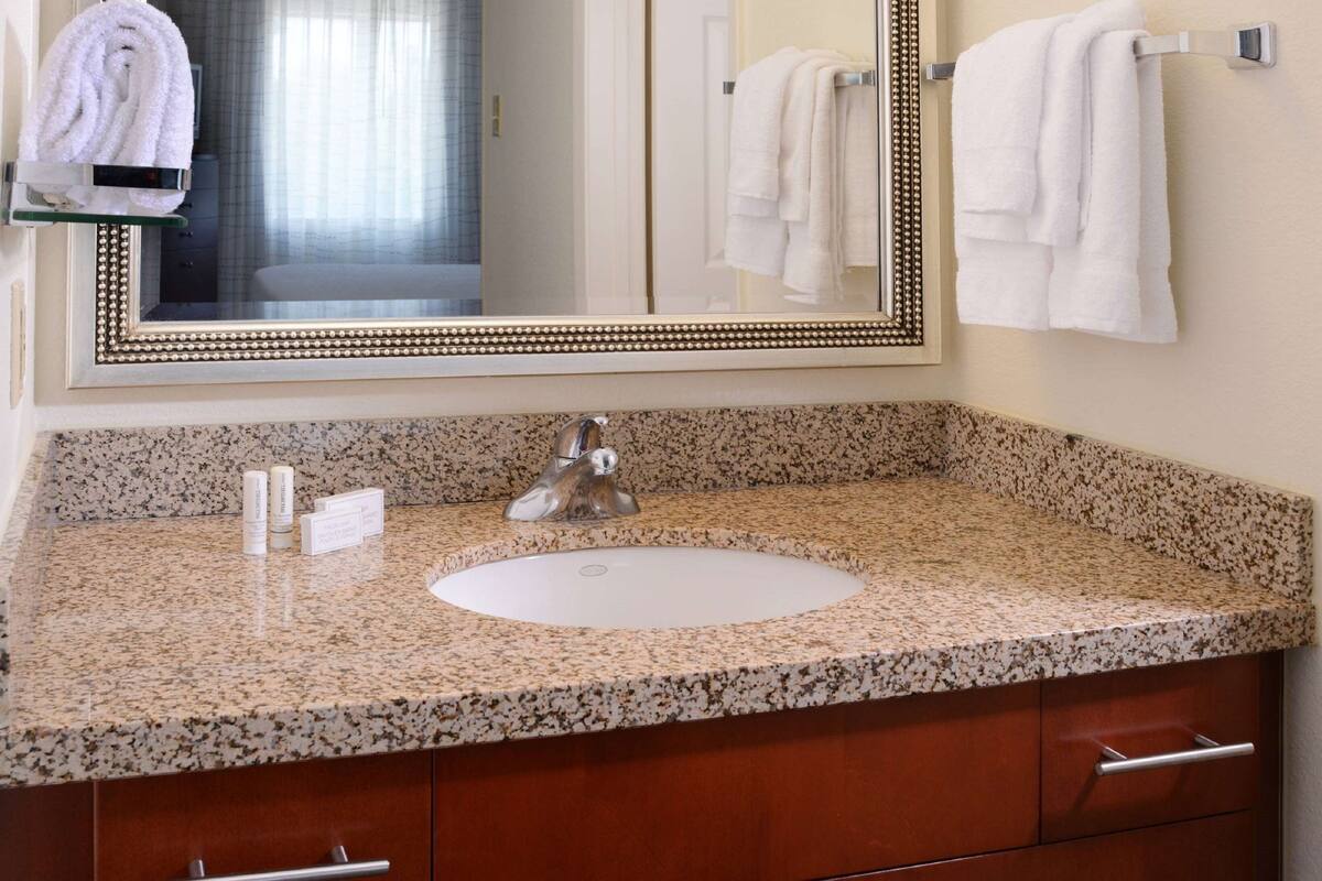 A bathroom with a granite countertop, a white sink, a mirror with a decorative frame, and a towel rack with folded towels.