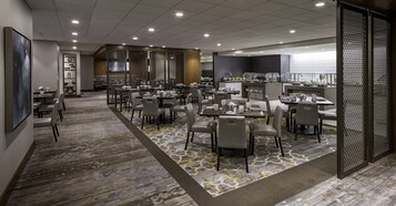A hotel dining area with multiple tables and chairs, a kitchen in the background, and a patterned carpet.