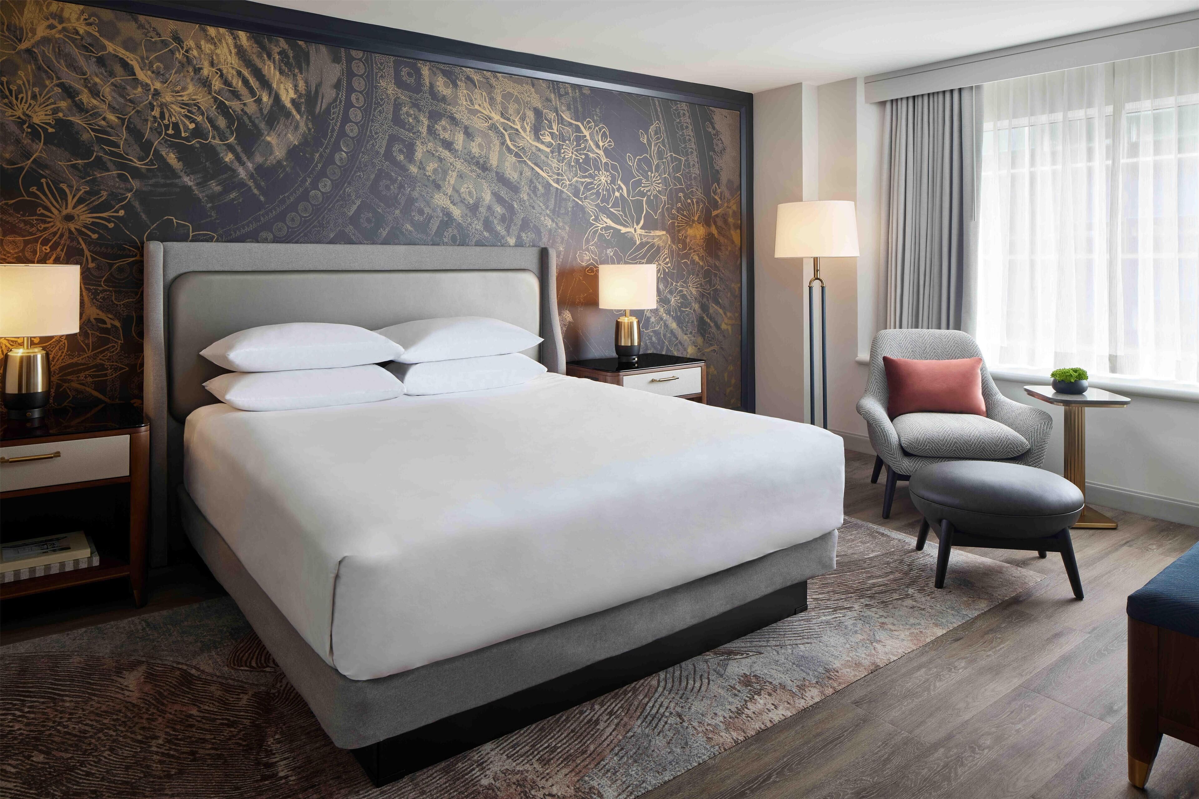 A modern hotel room with a large bed, two bedside tables with lamps, a chair, and a view of the city through sheer curtains.