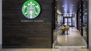 A Starbucks Reserve entrance with a logo on the wall, a reception area with a counter and baskets of pastries, and a modern interior with a wooden wall and glass doors. - Grand Hyatt Washington (Washington)